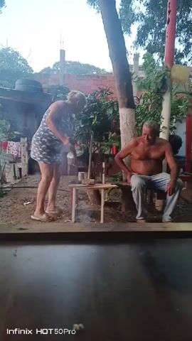The video shows two people outdoors near a small campfire. A woman on the left is standing and appears to be holding a metal object and interacting with a small table. A man on the right is sitting on a bench, shirtless, and also seems to be holding a metal object. The ground is covered in dirt and gravel, with some scattered leaves and debris. There are trees and foliage in the background, along with some structures. The lighting suggests it is daytime, possibly late afternoon or early evening. There is smoke rising from the campfire, indicating it is lit. The video is low resolution and has a watermark.