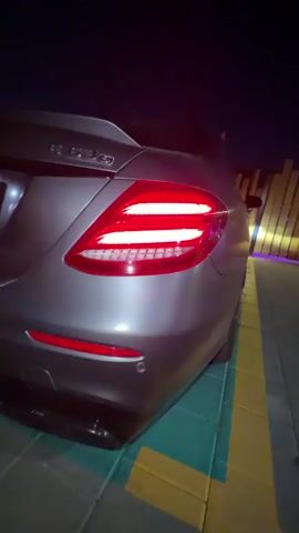 The video captures the rear of a silver Mercedes-Benz E-Class car at night. The taillights are illuminated in a bright red, with a distinctive horizontal light bar and accent lights. The car's emblem, appearing to be "E 350," is visible on the trunk. The car is parked on a paved surface with green and yellow markings, possibly in a parking lot or driveway. In the background, there is a structure with vertical elements illuminated by purple and yellow lights. The scene is dark, with the car's taillights being the primary light source. The camera is positioned low, focusing on the car's rear design.