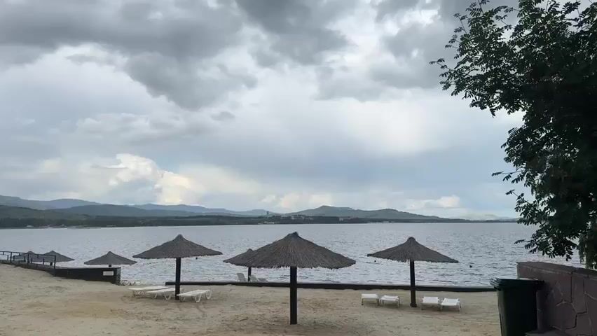 This media file depicts a serene beach scene at Yakty-Kul', Russia. The primary subjects are the sandy beach, the calm lake stretching towards a distant, verdant mountain range, and the overcast sky. Scattered across the beach are several thatched umbrellas providing shade, and a few empty lounge chairs suggest a peaceful, unoccupied setting.

The scene is set during what appears to be a cloudy day, likely late morning or early afternoon, given the diffused light. The weather is somber, with heavy, grey clouds dominating the sky, hinting at potential rain, but no precipitation is actively occurring. The mountains in the background are lush with green foliage, indicating a temperate climate. The water is still, with only gentle ripples disturbing its surface.

Notable details include a dark green trash bin on the right side of the frame, bordered by a stone wall, and a wooden walkway extending into the lake on the left. A few buoys are visible in the water, suggesting it is a designated swimming or recreational area. The overall mood is tranquil and contemplative, amplified by the lack of human presence and the subdued lighting. There is no visible text within the media file. The focus is on the natural landscape and the simple amenities of a lakeside resort.