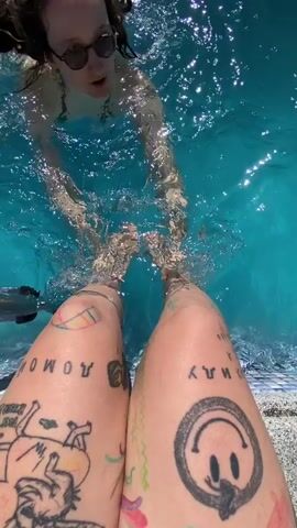 The video shows a first-person perspective of two legs covered in tattoos, standing on the edge of a swimming pool. In the background, a person with dreadlocks and sunglasses is in the bright blue water, smiling and reaching out to hold the feet of the person in the foreground. The water is clear and sparkling, suggesting a sunny day. The tattoos on the legs include a smiley face, a rainbow, and various other drawings and text. The person in the foreground is holding the feet of the person in the water, creating a playful and intimate moment. The scene evokes a sense of fun and relaxation on a warm day.