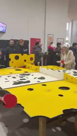 The video shows an indoor event with a crowd of people and a ping pong table decorated to look like Swiss cheese.  The camera first focuses on two people playing ping pong at the cheese-themed table. The table is painted yellow with large, round “holes” cut out to resemble cheese. The camera then pans up to show the crowd of people behind the table.  Pink lights and smoke can be seen on a stage in the distance, indicating a performance area. People are standing around, some watching the ping pong game and others moving towards the performance area.