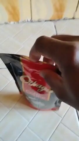 The video shows a person's hand reaching into a bag of coffee. The bag is mostly black with red and gold accents, and the word "Original" can be seen. The hand appears to be dark-skinned. The bag is placed on a surface with light beige tiles. After the hand reaches inside the bag, it pulls out and the bag is held up, revealing the dark coffee grounds inside. The hand then appears to be pinching the bag to close it. Finally, the hand releases the bag. Scissors can be seen in the background on the tiled surface. The tiles behind the bag are cracked.