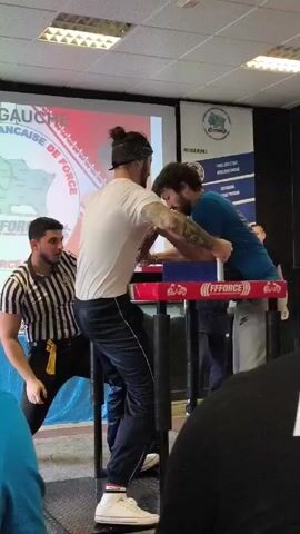 Here's a description of the video:

The video shows an arm wrestling match taking place indoors. Two men are engaged in a contest, their arms locked on an arm wrestling table. The man on the left is wearing a white t-shirt and dark pants. His hair is pulled up in a bun. The man on the right is wearing a blue t-shirt and light grey sweatpants. A referee in a black and white striped shirt oversees the match, watching intently. Several spectators are visible around the table. Banners in the background bear the names of the event and sponsoring organizations. The room is brightly lit with fluorescent lights.