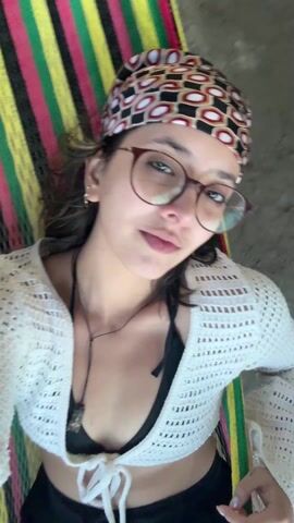The video starts with a close-up of a young woman lying in a colorful hammock. She has on glasses, a patterned headscarf, a black bikini top, and a white, loosely knitted cardigan. She looks directly at the camera with a slight smile. The camera then quickly tilts upwards and pans to show a beach scene. A man is visible in the distance, sitting in a chair on the sandy beach near the ocean. The background shows a cloudy sky over the ocean. The camera angle implies the woman is beneath a thatched roof, looking out at the beach.