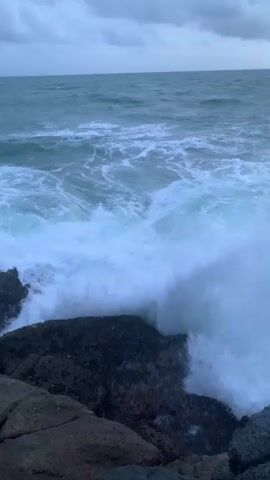 A video clip of waves crashing against rocks on a cloudy day. The water is choppy and appears to be a deep blue-green color, with white foam from the waves. The sky is overcast with dark clouds, and the light is dim. The rocks in the foreground are dark and rough. The scene is serene and powerful, conveying a sense of the raw beauty of nature.
