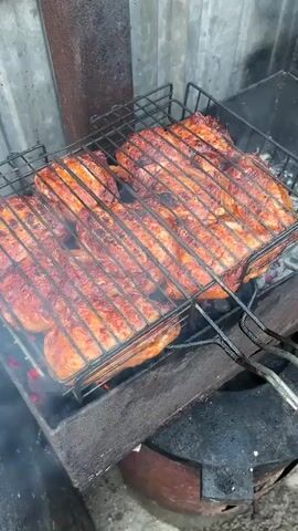 The video shows several pieces of meat on a grill over hot coals. The meat appears to be cooking and has grill marks on it. Smoke is rising from the grill. The grill is black metal and sits on top of a dark brown object, possibly a barrel or a stand. In the background, a corrugated metal wall is visible. The overall environment suggests an outdoor cooking area.
