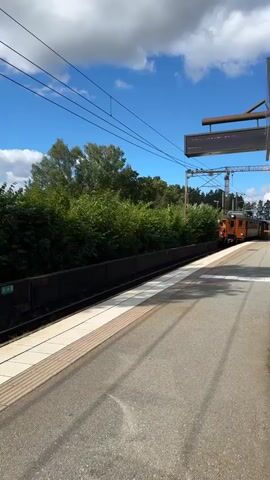 An orange and brown train passes by on a railway track. The train is moving from left to right across the screen. The train has multiple windows and appears to be an older model. In the background, there are trees and blue sky with white clouds. The train is on a track next to a platform that has a textured surface. Overhead, there are power lines and a sign.