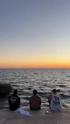 Three men sit facing a body of water with the sky transitioning from light blue to orange at the horizon, indicating sunset. The man on the left wears a black t-shirt with a graphic design on the back. The middle man is shirtless. The man on the right wears a white tank top with the words "Jiu Jitsu" printed on it. A white towel is placed on the ground to the left of the men, and a smartphone is on the ground to the right of the middle man. The water is rippled, suggesting light waves. A small crescent moon is visible in the upper left quadrant of the sky.
