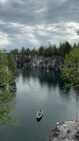 This video shows a panoramic view of a quarry with a lake. The quarry walls are made of marble and are very tall and steep. There are trees growing on top of the quarry walls. The lake is dark blue and reflects the sky and the quarry walls. There are several boats on the lake. One boat is in the foreground and has two people in it. Other boats are in the background and also appear to have people in them. The sky is cloudy and the lighting is somewhat dim, suggesting an overcast day. The camera is positioned above the lake, looking down at the scene.