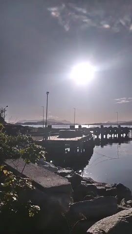 A vertical shot of a calm body of water reflecting the bright sun and a clear sky. A pier with pilings extends into the water on the left, with a small boat moored nearby. In the distance, hills or mountains form a distant horizon. The foreground features large rocks and some vegetation. A yellow buoy is visible in the water further out.