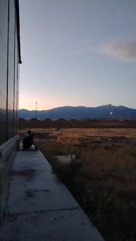 The video shows an outdoor scene at dusk. In the foreground, a dark, reflective building wall occupies the left side of the frame. Adjacent to the building is a concrete walkway, with two individuals crouching on it. Beyond the walkway, a wide expanse of dry, brown grass and dirt stretches into the distance. In the middle ground, there are several mounds of earth, possibly construction sites. Further back, a range of blue-hued mountains forms the horizon. The sky is a soft gradient of blue and gray, with a faint, wispy cloud. The overall impression is one of a rural or developing area at the end of the day.