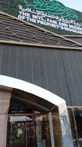 The video shows the exterior of "The Intl. Fair and Museum of the Prophet's Biography". The building has a modern design with a facade featuring a large, angled gray surface and a green sign with Arabic script. Below the gray surface is a row of windows reflecting the surrounding environment. There is a covered entrance with a stone frame. The ground in front of the building is tiled and includes a few small plants in planters. People can be seen walking inside the building.
