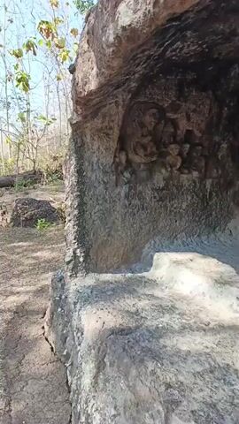 The video pans across an ancient rock carving within a cave. The carving depicts figures that appear to be religious or mythological in nature, with intricate details suggesting a historical or cultural significance. The cave itself is rough and natural, with uneven rock surfaces and a sandy floor. The surrounding environment is outdoors, with trees and foliage visible in the background, indicating a natural, possibly remote, setting. Sunlight filters into the cave, illuminating the carvings and the interior. The camera movement is steady, allowing for a clear view of the carvings and the rock formations.