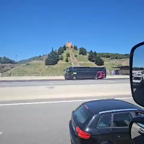 The video shows a highway scene in daylight. A black bus with a pink logo drives on the highway, and the camera, positioned within another vehicle, pans upward to reveal a large, terraced hill. A long, stone staircase ascends the hill, culminating in a tall, reddish-brown structure resembling a simplified castle at the summit. Trees and shrubbery dot the hillside. The sky is clear and blue. A black car is visible in the foreground, and a white car drives alongside the bus.