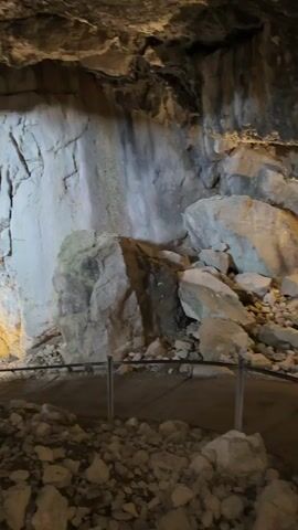 The video shows the interior of a cave with rough, rocky walls and ceiling. A path with a metal railing runs through the foreground, surrounded by piles of rocks. Several people are walking along the path, heading away from the camera. The cave is illuminated with artificial lighting, creating shadows and highlighting the texture of the rock.