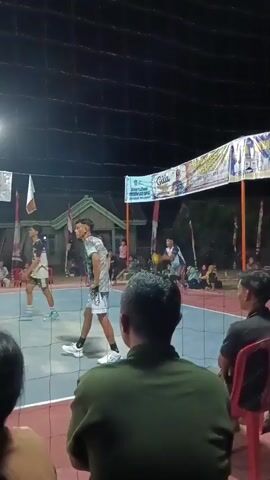 The video shows a volleyball game at night. The court is illuminated by overhead lights. Spectators are visible in the foreground, watching the game. On the court, players are in action. One player in a blue and yellow jersey is jumping to spike the ball, while another player in a black and white jersey is blocking. Several other players are positioned around the court. Banners are hanging in the background above the court. The overall scene depicts a competitive sports event taking place outdoors in the evening.