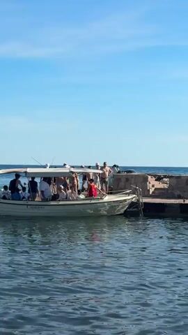 A small white boat with a canopy is docked next to a stone pier. Several people are on the boat, and some are standing at the edge, appearing to be ready to jump into the water. The water is calm and blue, and the sky is clear. In the distance, a large cruise ship is visible on the horizon. A few buoys are floating in the water. The overall scene depicts a sunny day with people enjoying a day out on the water.