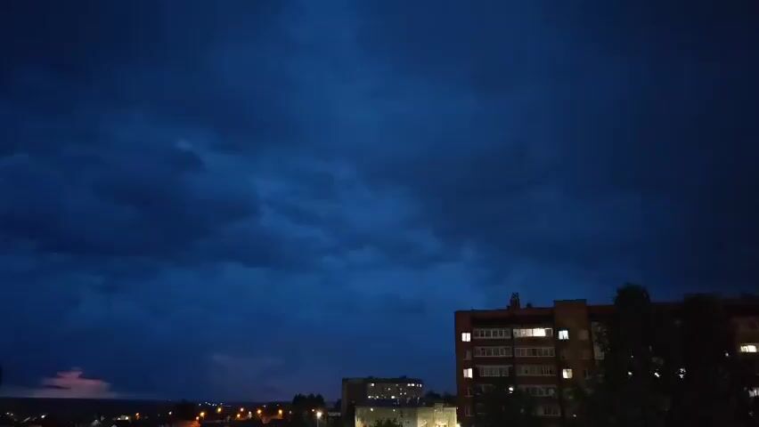 A city skyline is visible at dusk or night, with dark, stormy clouds dominating the sky. A few buildings are visible, including a multi-story apartment building on the right with some lights on. There is a faint pinkish glow on the horizon in the distance, possibly from lightning or a distant city. Trees are visible in the foreground on the right.