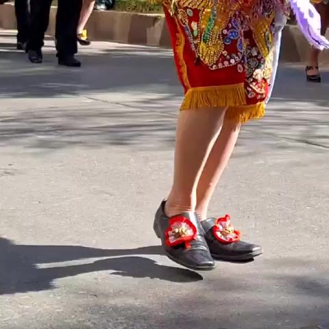 The video shows a close-up of a person's legs and feet dancing outdoors on a paved surface. The person is wearing traditional, elaborately decorated red pants with gold trim and fringe, and black shoes adorned with red and gold floral decorations. The person's legs are in motion, indicating they are dancing. The background is slightly blurred, showing hints of other people and foliage. The lighting suggests daytime.