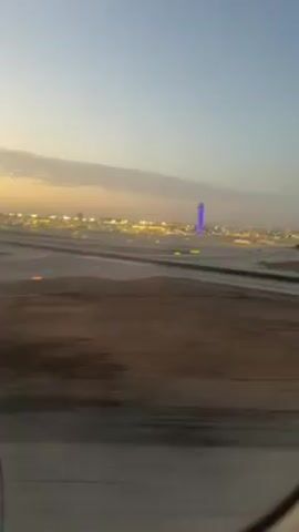 The video, shot from an airplane window, shows an airport runway at what seems like dusk or dawn. In the distance, a tall, illuminated tower with a blue top can be seen, surrounded by other airport buildings and lights. The runway has distinct markings and is bordered by dirt. The sky is a gradient of light blue to pale yellow, indicating either sunrise or sunset. The plane appears to be moving along the runway.