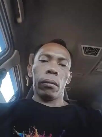 The video shows a close-up, selfie-style shot of a man's face. He appears to be sitting in the driver's seat of a car, as indicated by the car's interior visible in the background, including air vents and parts of the car's ceiling. The man is wearing a black t-shirt with a colorful, abstract design on the front. His facial expression is neutral, and he is looking directly at the camera. The lighting suggests it is daytime, possibly outdoors or in a well-lit area. No other people or significant actions are depicted.