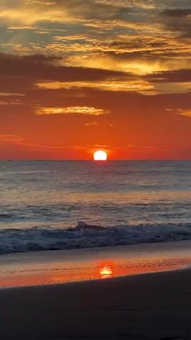 A serene sunrise paints the sky with vibrant hues of orange and yellow. The sun, a glowing orb, is positioned at the horizon, casting a warm reflection on the calm sea. Gentle waves lap against the sandy shore, creating a peaceful and tranquil atmosphere. The clouds, illuminated by the rising sun, add depth and texture to the breathtaking panorama.