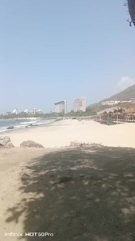 The video shows a sandy beach on a sunny day. The beach is bordered by a treed area and some large rocks in the foreground, while in the distance there are several high-rise buildings, some smaller buildings, and a mountain. The beach itself has some straw umbrellas and a few trash cans. There are some waves gently breaking on the shore. The sky is clear and blue. The camera is held at a relatively low angle and moves slowly.