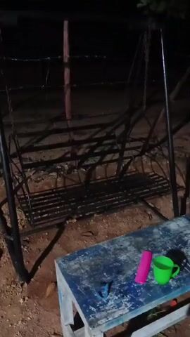 The video shows a static shot of an empty metal swing set in an outdoor setting at night. A wooden table is in the foreground, with a pink object, a blue object, and a green mug on it. The ground appears to be dirt and pebbles. Barbed wire is visible in the background, suggesting a fence. Shadows are cast across the scene, likely from lighting sources not visible in the frame.