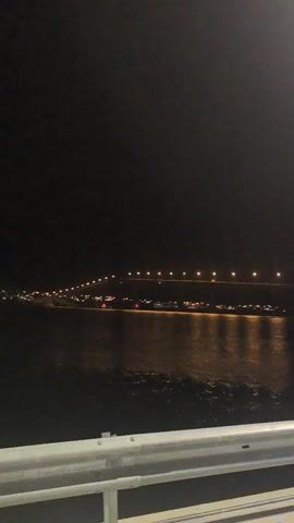 The video shows a night view of a bridge over water. The scene is dark, with the only light sources being the bridge lights and reflections in the water. The camera is positioned behind a highway guardrail. There is little detail visible beyond the bridge and water.