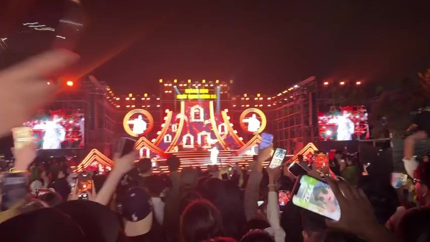 The video captures an outdoor night concert in Măng Đen, Vietnam. A male performer, dressed in a long white coat, is on an elaborate, multi-level stage, singing into a microphone. The stage is brightly lit with numerous red and white LED lights, forming intricate geometric patterns and highlighting golden-framed decorative elements resembling mirrors or screens. Large LED screens behind and to the sides of the performer display abstract visuals and close-ups of him.

A large crowd of people is gathered in front of the stage, facing the performer. Many attendees are holding up their cell phones to record the event, while others are cheering and waving. The atmosphere appears lively and energetic.

Visible text on the main LED screen reads "MĂNG ĐEN KHÁT VỌNG VƯƠN XA". The scene is set at night, under clear conditions, emphasizing the stage's lighting and screen displays. Musical instruments, including drums and a guitar, can be briefly seen on the stage behind the performer.