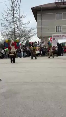 The video shows a group of people in traditional costumes dancing in a town square in front of a crowd. The dancers wear furry costumes and large, colorful headdresses. Some appear to be wearing masks. Some also wear bells on their waistlines. A few people in the crowd are in traditional folk dress, while others are in modern street clothing. A banner says “Chestit Praznik, Aidemir” in Cyrillic script. The backdrop is a plain grey building with a small Bulgarian flag on display. A tent is set up next to the building. The dancers are arranged in a circle, and people play musical instruments while others dance. The square is paved with asphalt.