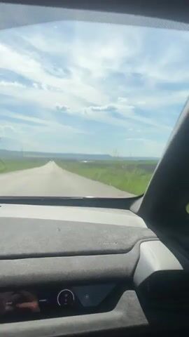 The video captures a first-person view from inside a car driving on a straight, paved road through a rural landscape. The interior features a dashboard with a suede-like finish, a digital instrument cluster displaying speed, and a central infotainment screen with navigation.

A driver's gloved hand is visible on the steering wheel, indicating active driving. The car's speed changes from 107 km/h to 113 km/h throughout the clip, and the instrument cluster also shows "SPORT" mode and a "HYBRID" indicator. The infotainment screen displays a map with a route, showing "2.6 km" remaining, and "AUTO 20.0 °C". A "Lamborghini" logo is faintly visible on the bezel of the infotainment screen.

Outside the car, the scene is dominated by vast green and yellow fields stretching to the horizon under a partly cloudy blue sky. Several power lines and transmission towers are visible in the distance on both sides of the road. The sunlight suggests it is daytime. The yellow fields likely indicate blooming rapeseed or similar crops. The overall impression is of a fast drive through an open, agricultural area. The audio features prominent engine sounds and wind noise, consistent with a vehicle moving at speed.