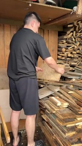 A man wearing a black t-shirt and black shorts is standing in what appears to be a shed or workshop. He is holding a piece of wood in his hands, examining it. In the background, there are stacks of lumber and other wooden materials. The man is standing near a pile of wood scraps. He moves the piece of wood around as he inspects it.