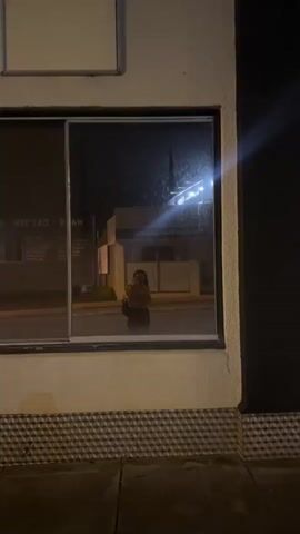 The video opens at night, capturing a person's reflection in a large storefront window. The individual is holding a phone, seemingly recording or taking a selfie. Reversed text, including "DAY SPA" and "HAIR," is visible on the window, suggesting the type of business. The reflection also shows glimpses of a street with lights. The camera then transitions to an outdoor view of a street at night. A prominent, illuminated McDonald's sign, featuring the golden arches, is visible on the left. Car headlights and streetlights create long, vibrant light streaks across the frame, indicating movement, possibly from a car or the person walking. Several cars are seen on the road. The setting appears to be an urban or suburban area after dark.