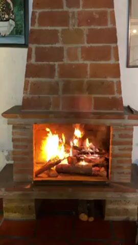 The video shows a fireplace made of bricks. A fire is burning inside the fireplace. The fireplace has a traditional design, with a hearth, a firebox, and a chimney. The fire is burning brightly, with flames reaching up towards the top of the firebox. Several logs are burning in the fireplace. The floor in front of the fireplace is also made of bricks. There are two framed images on the white walls to the left and right of the fireplace.