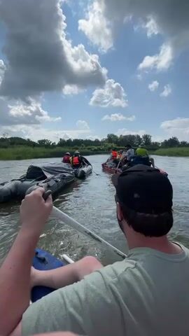 A group of people are on a river in rafts. One person in the foreground is paddling a raft with a paddle. There are three other rafts visible in front of them. The rafts are connected by ropes. The people in the rafts are wearing life jackets. The river is surrounded by green vegetation. The sky is blue with some white clouds.