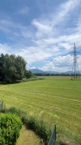 The video shows a landscape view with a large green field in the foreground. A tall metal power line tower stands in the middle ground, with power lines stretching across the sky. In the background, there are mountains and a blue sky with white clouds. The lighting suggests it is daytime, likely a sunny or partly cloudy day. There are no people or animals visible in the video. The camera pans slightly to the right, revealing more of the mountainous landscape.