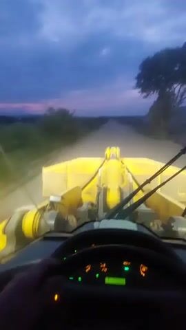The video shows a first-person view from inside a vehicle, likely a front-end loader, as it drives down a dirt road at dusk. The vehicle is yellow and has a large bucket attachment at the front. The dashboard lights are illuminated, with a speedometer indicating a speed of approximately 20. The sky is dark blue with some clouds. Vegetation is visible on either side of the road, and a tree is on the right. The headlights of the vehicle are on, illuminating the road ahead. The driver's hands are visible on the steering wheel. The camera is relatively stable, suggesting the vehicle is moving at a moderate pace. There is no audio in the video.