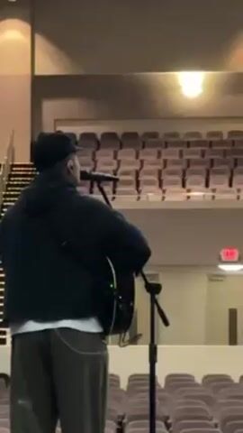 The video shows a male figure playing a guitar and singing into a microphone. He is seen from the back, standing on a stage in a large, empty auditorium. The auditorium is filled with rows of empty seats. The man is wearing a dark-colored hoodie, light-colored pants, and a baseball cap. A single light is visible in the ceiling, and an exit sign can be seen on the right side of the frame.