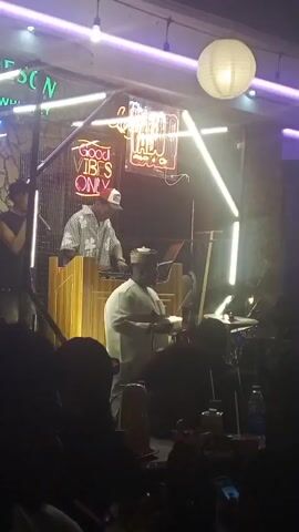 The video captures a vibrant night-time scene inside a venue in Lokoja, Nigeria, likely a bar or club, featuring a live musical performance. On a raised stage area, at least three performers are visible. In the center, a person wearing a light-colored, possibly traditional, outfit and a cap is actively playing a drum kit. To their left, a DJ in a checkered shirt and a cap stands behind a wooden console, seemingly mixing music. Another individual in a dark top is positioned to the far left, near a microphone, possibly singing or interacting with the audience.

The stage and background are brightly lit with various artificial lights, including white and purple/blue fluorescent tubes arranged geometrically. Several neon signs are prominent: "JAMESON IRISH WHISKEY" is partially visible on the upper left, and "GOOD VIBES ONLY" is clearly displayed in the center. Another red neon sign with cursive script is also present but unreadable. A white paper lantern light hangs from the ceiling above the stage.

In the foreground, a silhouetted crowd of people is seated at tables, observing the performance. Some audience members are seen holding or looking at their mobile phones. The overall atmosphere appears lively and engaging, with music playing and people enjoying the event.