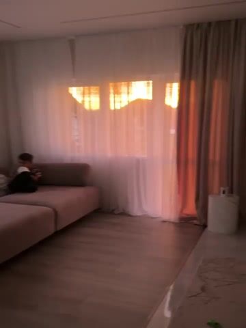 The video shows a living room with large windows covered by sheer white curtains and a darker beige curtain on the right. The setting sun casts a warm, orange glow through the windows, creating a silhouette of buildings and the sky. A pink sectional sofa is visible on the left, with a child sitting on it, looking towards the window. The floor is made of light-colored wood planks. A small white side table with a marble-like top is positioned near the right curtain. The overall ambiance is peaceful and serene, with the natural light dominating the scene.