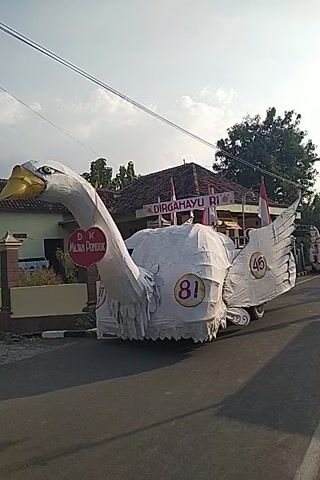 A large white swan-shaped float is parked on the side of a road. The float is decorated with banners that say DIRGAHAYU RI and feature the numbers 45 and 81. The swan's head is extended forward and its beak is yellow. A red and white Indonesian flag is attached to the float. The background shows houses and trees, and the sky is cloudy. The video is filmed vertically and is slightly tilted.