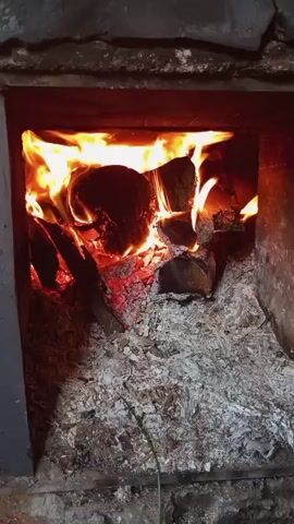 The video shows a wood-burning oven or fireplace in close-up.  Flames dance around several logs, illuminating them with bright orange and red light. The bottom of the oven is filled with a thick layer of grey ash. The structure appears to be made of stone or concrete blocks.  There is no visible movement except for the flames.