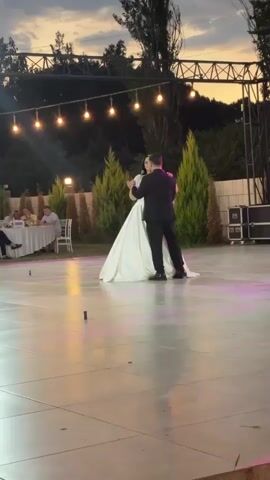 A man in a black suit and a woman in a white wedding dress are dancing on a tiled outdoor dance floor. The woman is wearing a veil. String lights are hung above the dance floor, and there are trees and a hillside in the background. To the left of the dance floor, there are tables with people seated, and to the right, there are speakers and equipment. The sky appears to be dusk.