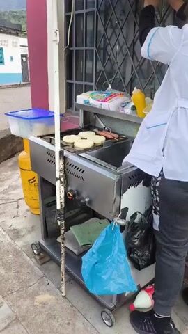 The scene shows a food cart on a sidewalk. A person in a white cook's jacket is working at the cart, which has a flat-top grill where four round arepas and two sausages are being cooked. The cart is made of stainless steel and has a lower shelf holding a green cloth and a blue plastic bag. A yellow propane tank is next to the cart. Various supplies, including napkins and a bottle of mustard, are on the cart's upper shelf. The background includes a building with a window behind the cart and a street with buildings and trees in the distance. The lighting is natural daylight.