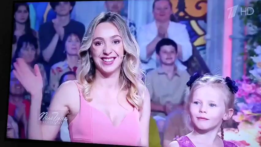 The video shows a television screen displaying a segment from a show. In the foreground, a young blonde woman in a pink dress is smiling and waving. To her right, a young blonde girl in a purple dress with a dark bow in her hair looks off to the side. In the background, an audience is visible, some people are clapping. The logo "1 HD" is visible in the upper right corner of the screen. The words "Наше Здесь" are visible on the screen in the lower left corner. The lighting appears to be bright and stage-like.