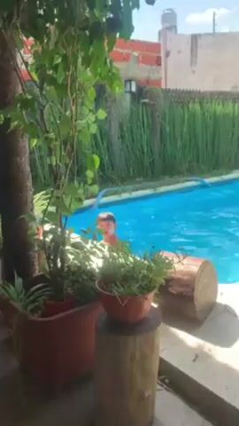 The video shows a sunny backyard scene with a swimming pool.  A boy is in the pool, followed by a panning shot to two girls near the edge. The pool is surrounded by a concrete deck and grass, with a fence and trees in the background.  Several potted plants are in the foreground. There are chairs, a table, and an umbrella. Clothes are drying on a clothesline.