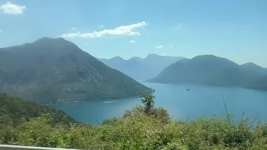 The video shows a scenic view of a bay surrounded by lush green mountains under a clear blue sky. The camera pans slowly, revealing the expansive blue water, with a few boats visible in the distance. The foreground is filled with dense green foliage, including trees and bushes, suggesting a natural, undeveloped landscape. The overall impression is one of serene natural beauty.