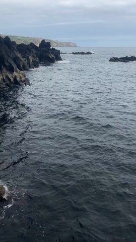 The video shows a rocky coastline on an overcast day. The sea is dark and choppy, with small waves lapping against the black volcanic rocks.  A few larger rock formations are visible in the distance. The sky is a muted grey.  There are no people visible in the shot.
