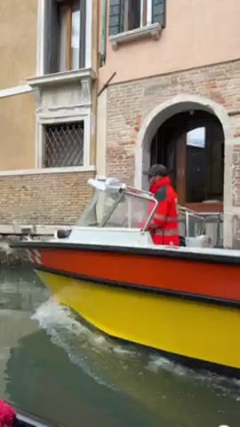 A yellow and orange ambulance boat passes down a narrow canal in Venice, Italy. Two people wearing red emergency service jackets are on the boat, one driving, the other as passenger. The boat has the words "AMBULANZA", "CROCE VERDE", "ANPAS", "118" and "Donazione di Marinette" on its side. The license plate reads "RV06949". Buildings line both sides of the canal. A gondola is partially visible in the foreground.